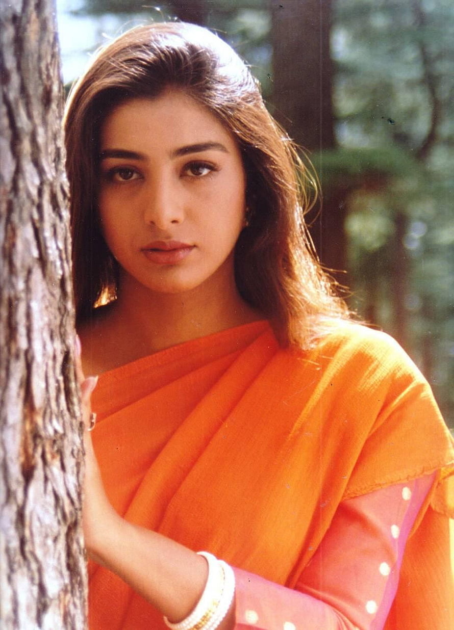 Tabu in a vibrant orange sari posing beside a tree trunk in a sunlit forest setting HD phone wallpaper