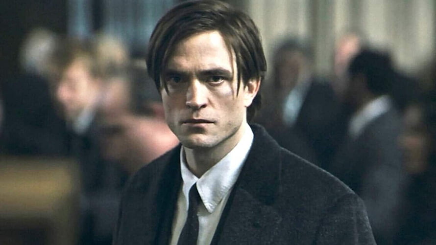 Robert Pattinson as Bruce Wayne in a dark coat with a serious expression in a crowded room HD wallpaper