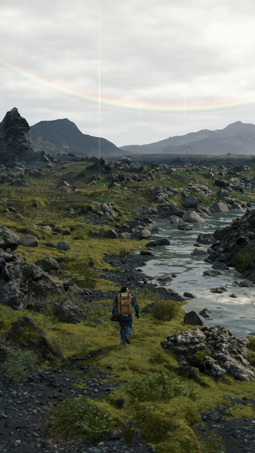 Death Stranding character walking through mossy volcanic landscape with river and rainbow HD phone wallpaper