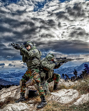 Two soldiers in camouflage tactical gear aiming rifles on a rocky mountain ridge under a dramatic cloudy sky HD phone wallpaper