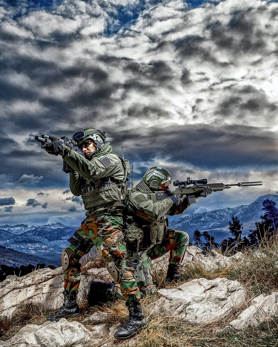 Two soldiers in camouflage tactical gear aiming rifles on a rocky mountain ridge under a dramatic cloudy sky HD phone wallpaper