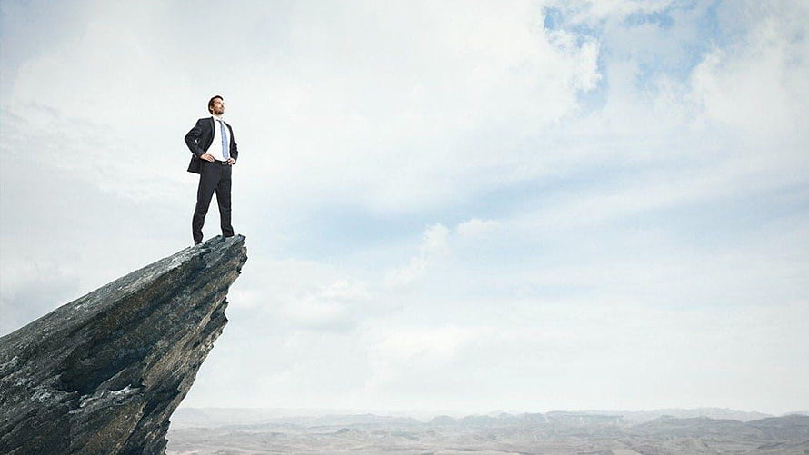 Businessman standing on a rocky cliff edge overlooking a vast landscape under a cloudy sky HD wallpaper