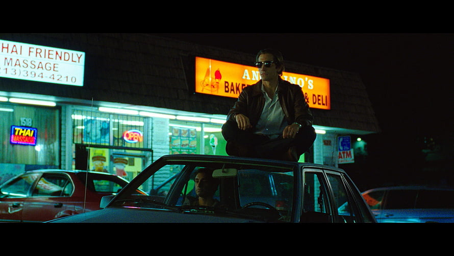 Nightcrawler Movie Scene: Man Sitting on Car Roof at Night in Front of Neon-Lit Bakery and Massage Shop HD wallpaper