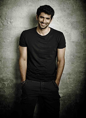 Aditya Roy Kapur smiling in a black t-shirt against a textured grey wall HD phone wallpaper