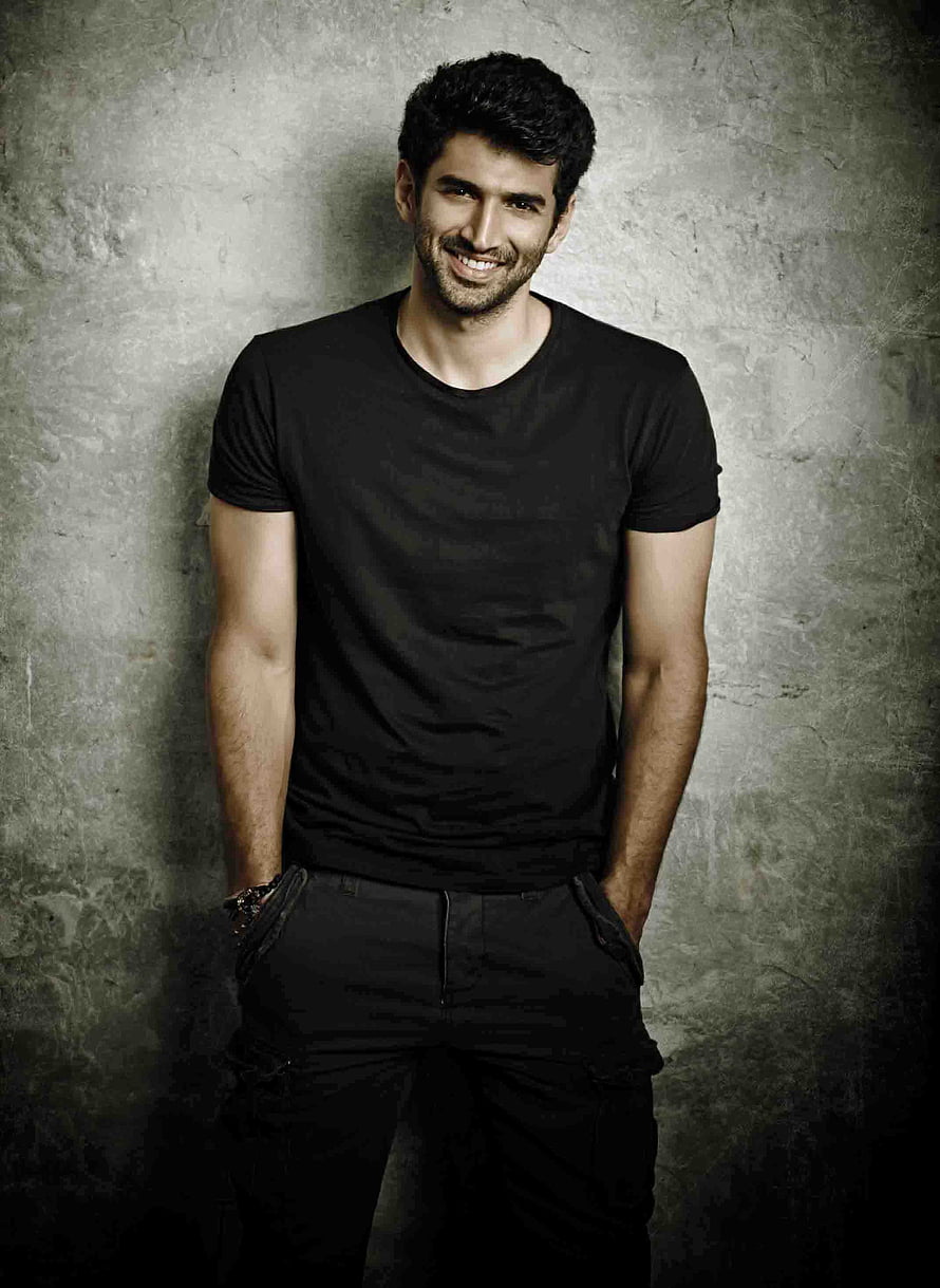 Aditya Roy Kapur smiling in a black t-shirt against a textured grey wall HD phone wallpaper