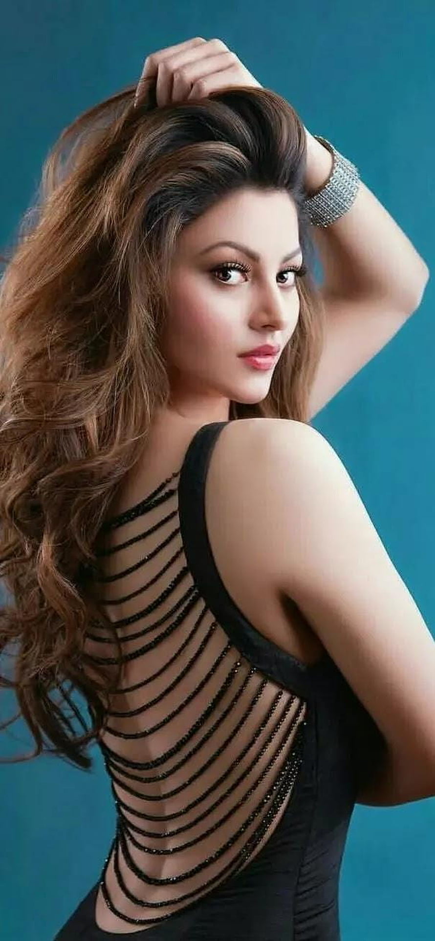 Urvashi Rautela in a black strappy back dress with long wavy hair against a teal studio background HD phone wallpaper