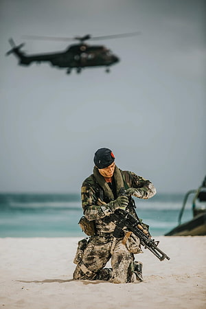 Special Forces Soldier Kneeling on Beach with Rifle and Helicopter in Background HD phone wallpaper