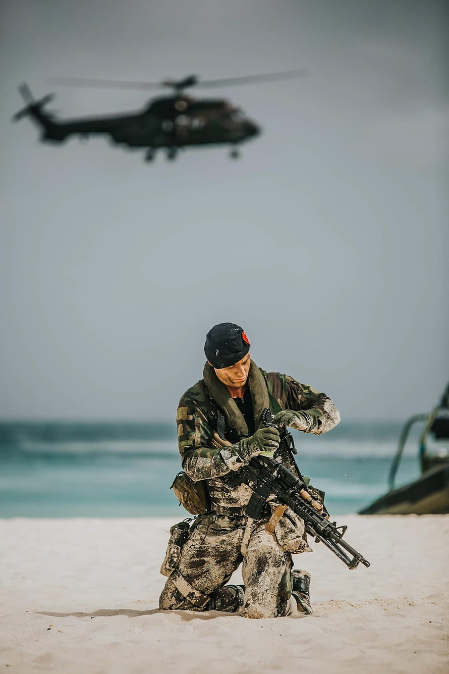 Special Forces Soldier Kneeling on Beach with Rifle and Helicopter in Background HD phone wallpaper