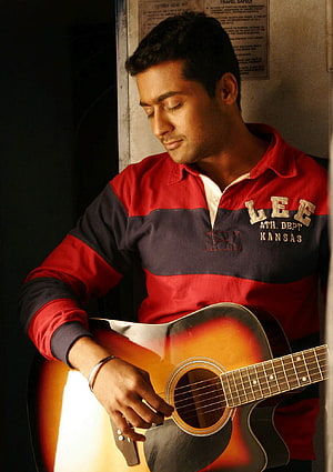 Surya playing an acoustic guitar in a red and navy polo shirt with warm cinematic lighting HD phone wallpaper