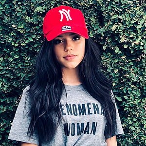 Jenna Ortega in a red New York Yankees cap and grey Phenomena Woman t-shirt against a green hedge HD phone wallpaper