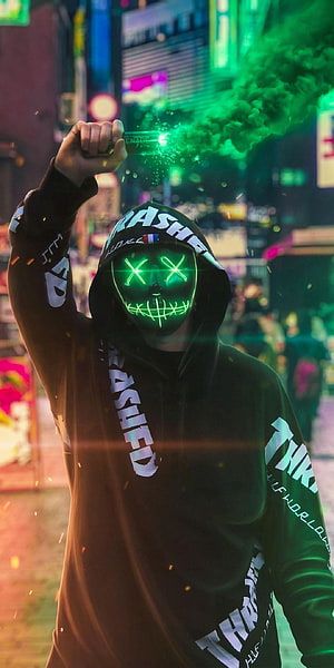 Person in black hoodie wearing a glowing green LED Purge mask holding a sparkler in a neon-lit city street at night HD phone wallpaper