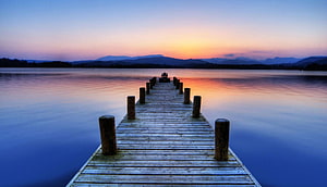 Serene wooden pier extending into a calm lake at sunset with distant mountains HD wallpaper
