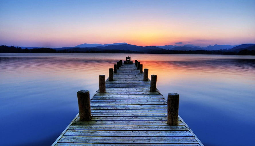 Serene wooden pier extending into a calm lake at sunset with distant mountains HD wallpaper