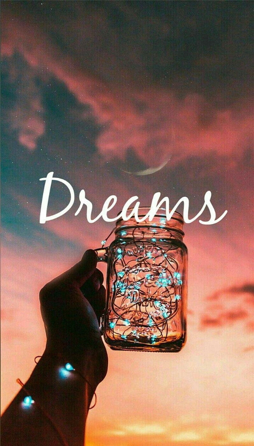 Hand Holding Glowing Fairy Light Jar Against Sunset Sky with Dreams Text HD phone wallpaper