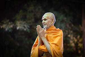Mahant Swami Ji in Orange Robes with Hands in Prayer Against Dark Foliage HD wallpaper