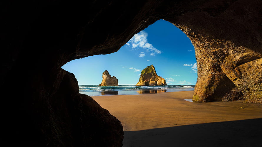 Coastal sea cave framing dramatic ocean rock formations and sandy beach under a clear blue sky HD wallpaper