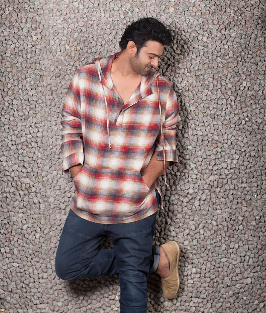 Prabhas in a red plaid hooded shirt and dark jeans leaning against a pebble wall HD phone wallpaper