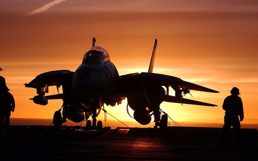 Silhouetted fighter jet on an aircraft carrier deck at sunset with crew members HD wallpaper