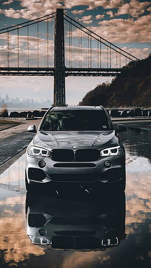 Silver BMW X5 M SUV Parked on Wet Pavement with Suspension Bridge and City Skyline in Background HD phone wallpaper