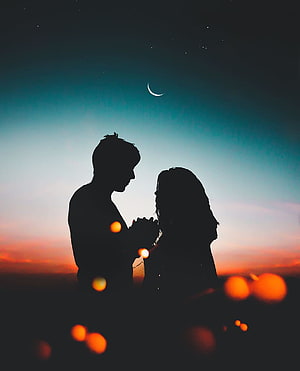 Silhouetted couple holding hands under a crescent moon at twilight with warm bokeh lights HD phone wallpaper