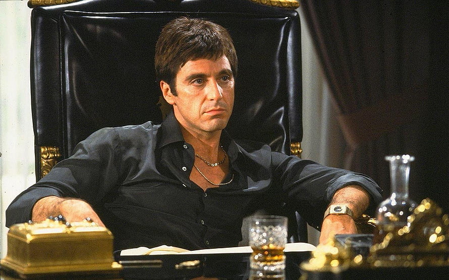 Tony Montana in Scarface seated in a black leather chair with gold desk accessories HD wallpaper