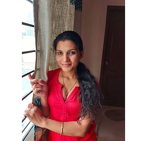 Resmi R Nair in a red top standing by a window with gold jewelry and long dark hair HD wallpaper
