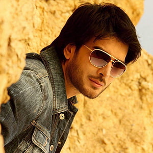 Stylish man in denim jacket and aviator sunglasses leaning against a textured stone wall HD phone wallpaper