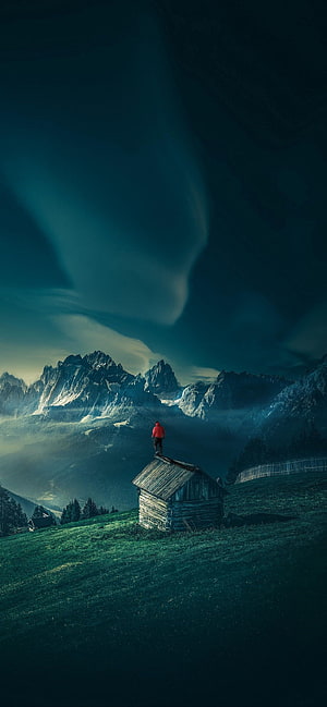 Lone Hiker on Alpine Cabin Roof with Dramatic Mountain Peaks and Misty Valley HD phone wallpaper