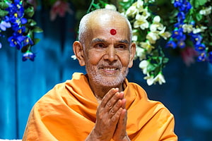 Mahant Swami Maharaj in saffron robes with hands pressed in prayer against a floral backdrop HD wallpaper