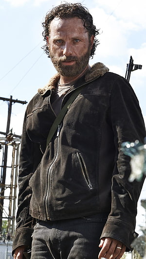 Rick Grimes in a worn leather jacket with a crossbow slung over his shoulder in a post-apocalyptic setting HD phone wallpaper
