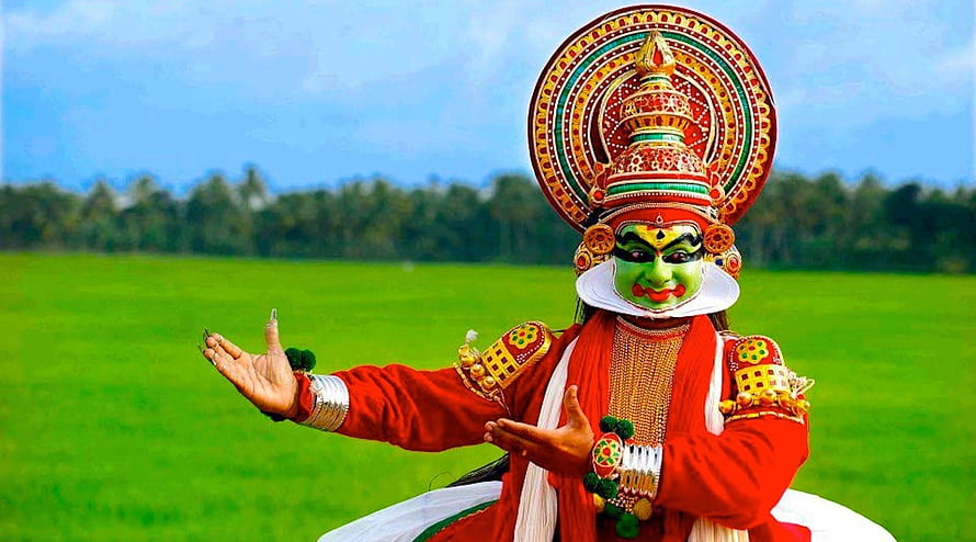 Traditional Kathakali dancer in vibrant red costume and ornate green face mask performing in a lush green field HD wallpaper