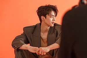Song Kang in a dark olive blazer against a warm orange studio backdrop HD wallpaper