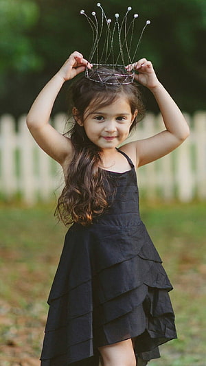 Little girl in a black tiered dress placing a wire crown on her head outdoors HD phone wallpaper