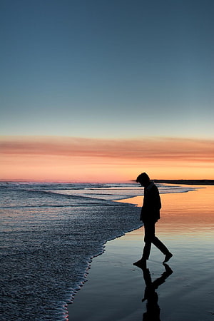 Lone figure walking along the shoreline at dusk with a warm orange horizon and calm ocean waves HD phone wallpaper