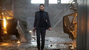 John Wick Walking Through Burning Wreckage in Dark Industrial Warehouse HD wallpaper