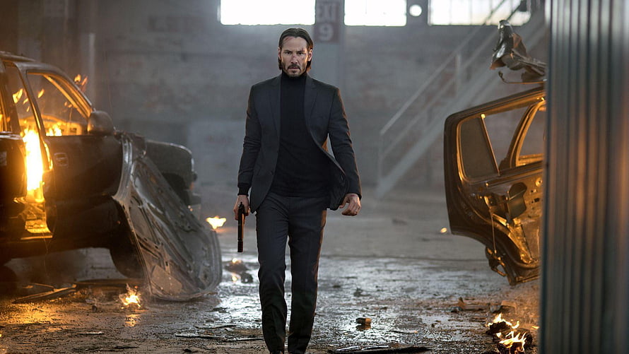 John Wick Walking Through Burning Wreckage in Dark Industrial Warehouse HD wallpaper