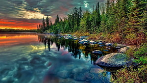 Vibrant sunset over a calm lake with evergreen forest and rocky shoreline HD wallpaper