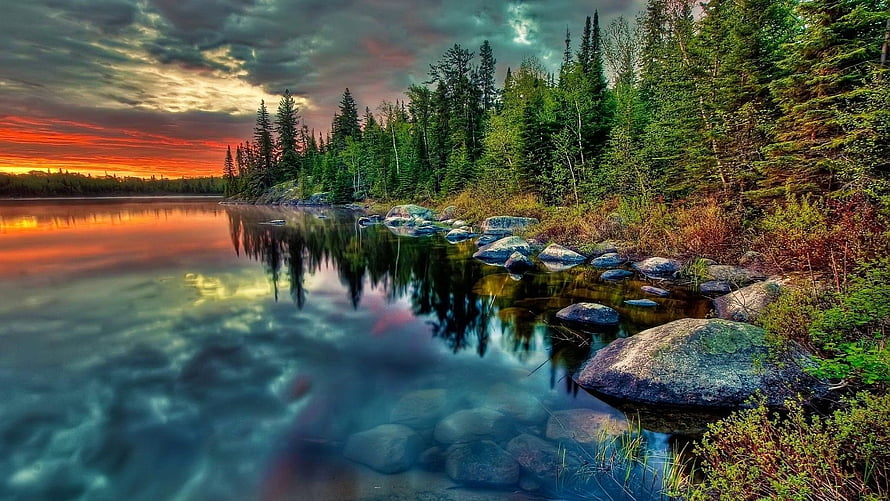 Vibrant sunset over a calm lake with evergreen forest and rocky shoreline HD wallpaper