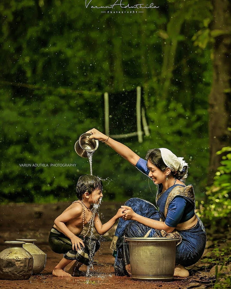 Indian mother pouring water over her son in a lush green forest setting HD phone wallpaper