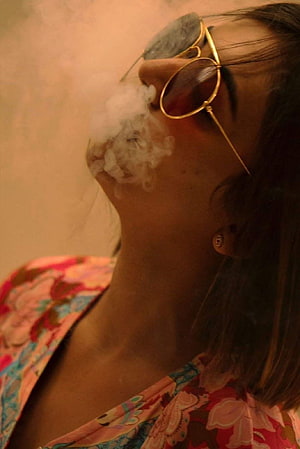 Woman in Gold Sunglasses Exhaling Smoke in Warm Ambient Light HD phone wallpaper