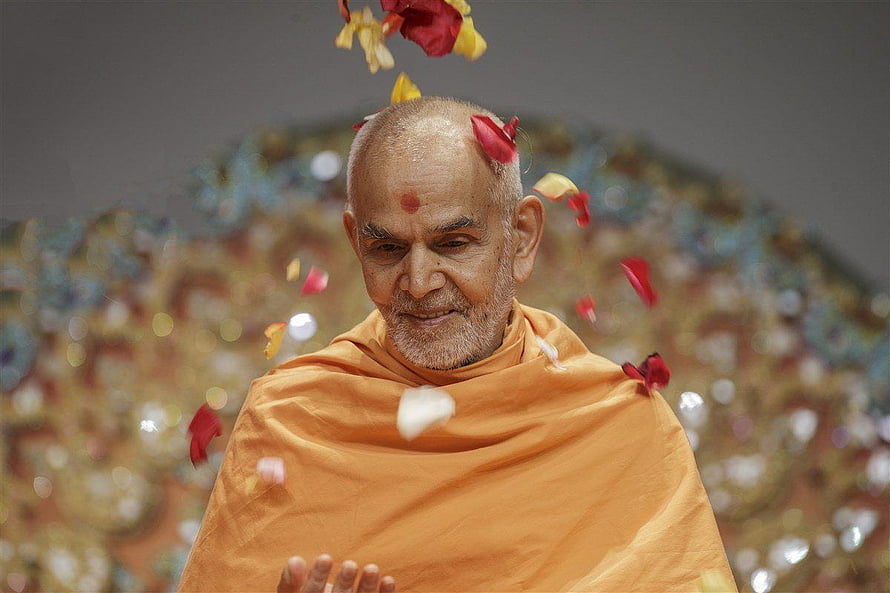 Mahant Swami Maharaj in saffron robes with falling flower petals and a serene expression HD wallpaper